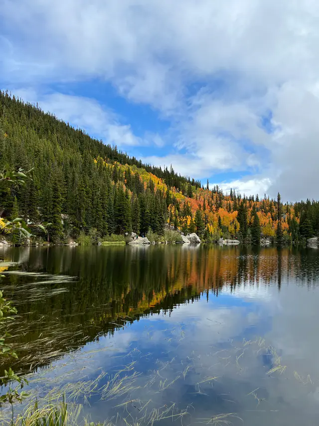 Autumn trees line a still alpine lake beneath a partly cloudy sky