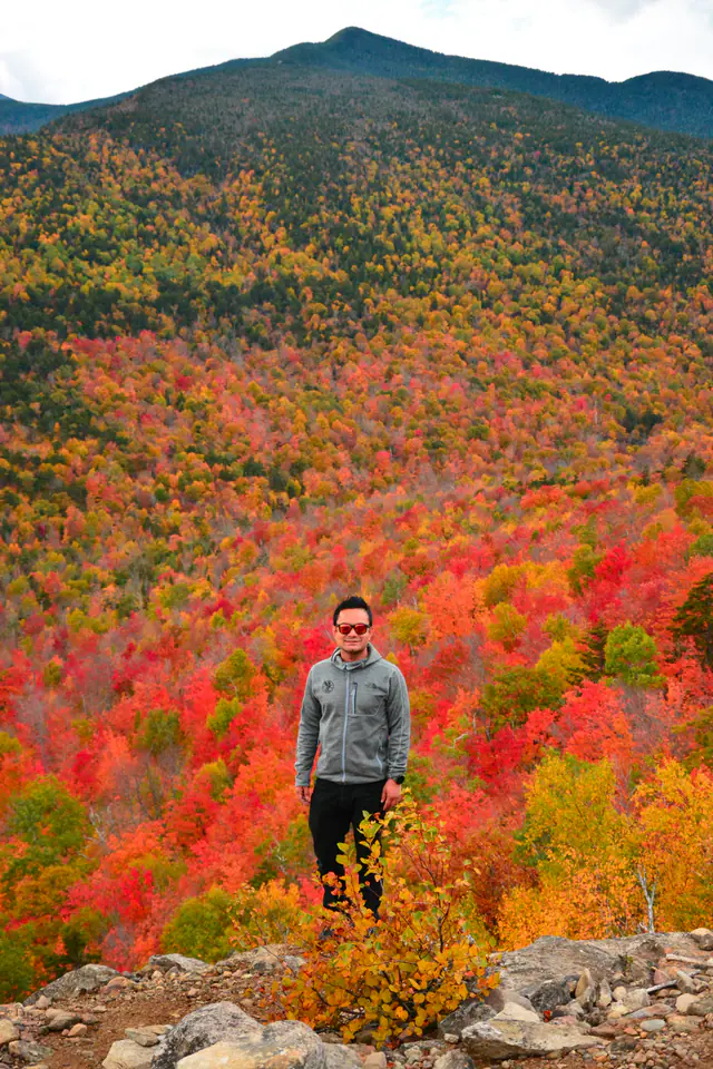 Nitesh stands before a mountain ridge covered in red, orange, and green autumn foliage