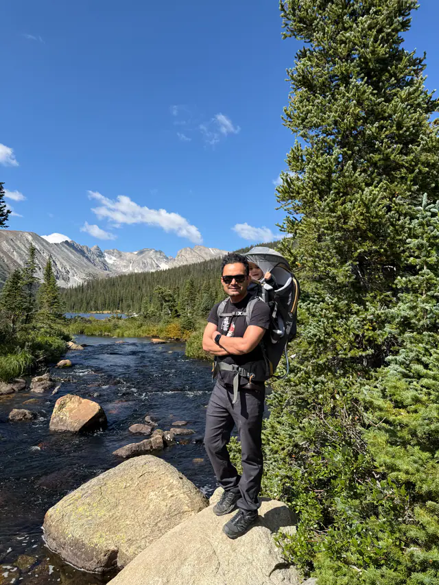 A hiker wearing a child carrier stands on boulders beside a mountain stream