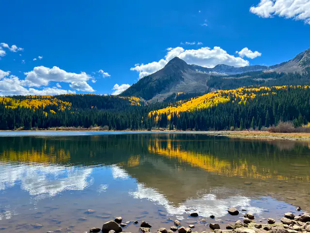A line of golden trees and a pointed peak reflect in a quiet lake