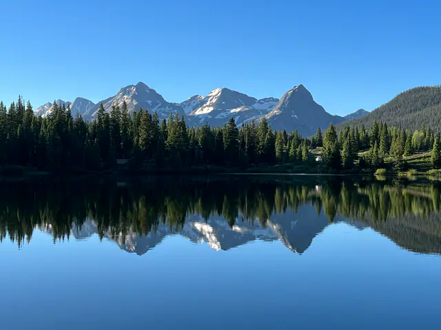 A row of mountain peaks and dark evergreens reflect in a calm lake