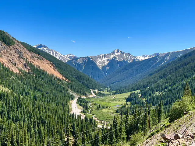 A green mountain valley and winding road stretch toward distant snowy peaks